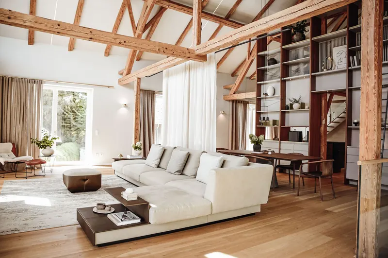 Das Wohnzimmer-Loft mit hellem Sofa, Nussbaumtisch, Leinenvorhängen und dem alten Dachstuhl.
